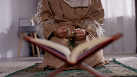 Nahaufnahme-Einer-Offenen-Kopie-Des-Korans-Auf-Einem-Ständer-Mit-Einer-Muslimischen-Frau,-Die-Im-Hintergrund-Mit-Gebetsperlen-Betet